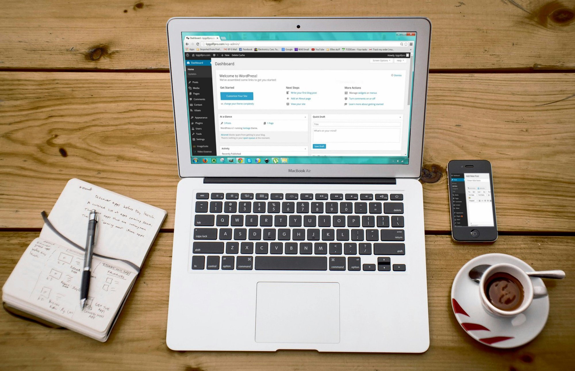 A MacBook Air displaying the WordPress dashboard sits on a wooden desk beside a handwritten notebook with a pen, a smartphone showing a WordPress screen, and a cup of coffee.